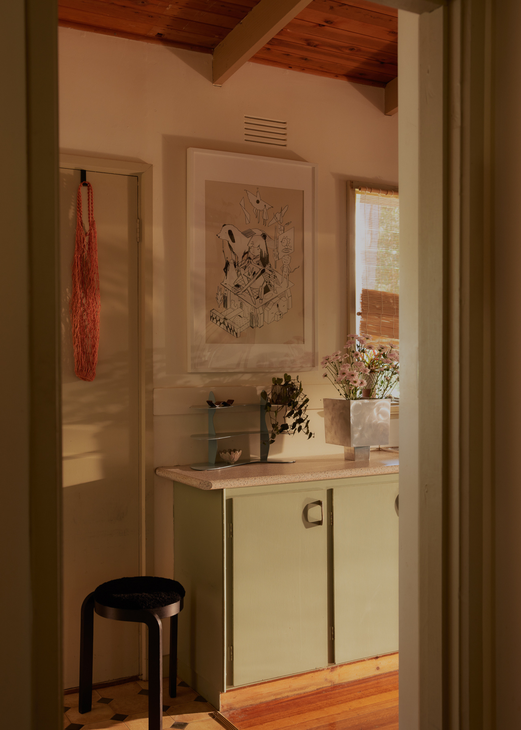 A kitchen with a small brown stool, sage green cabinets, framed art and a steel vase with flowers