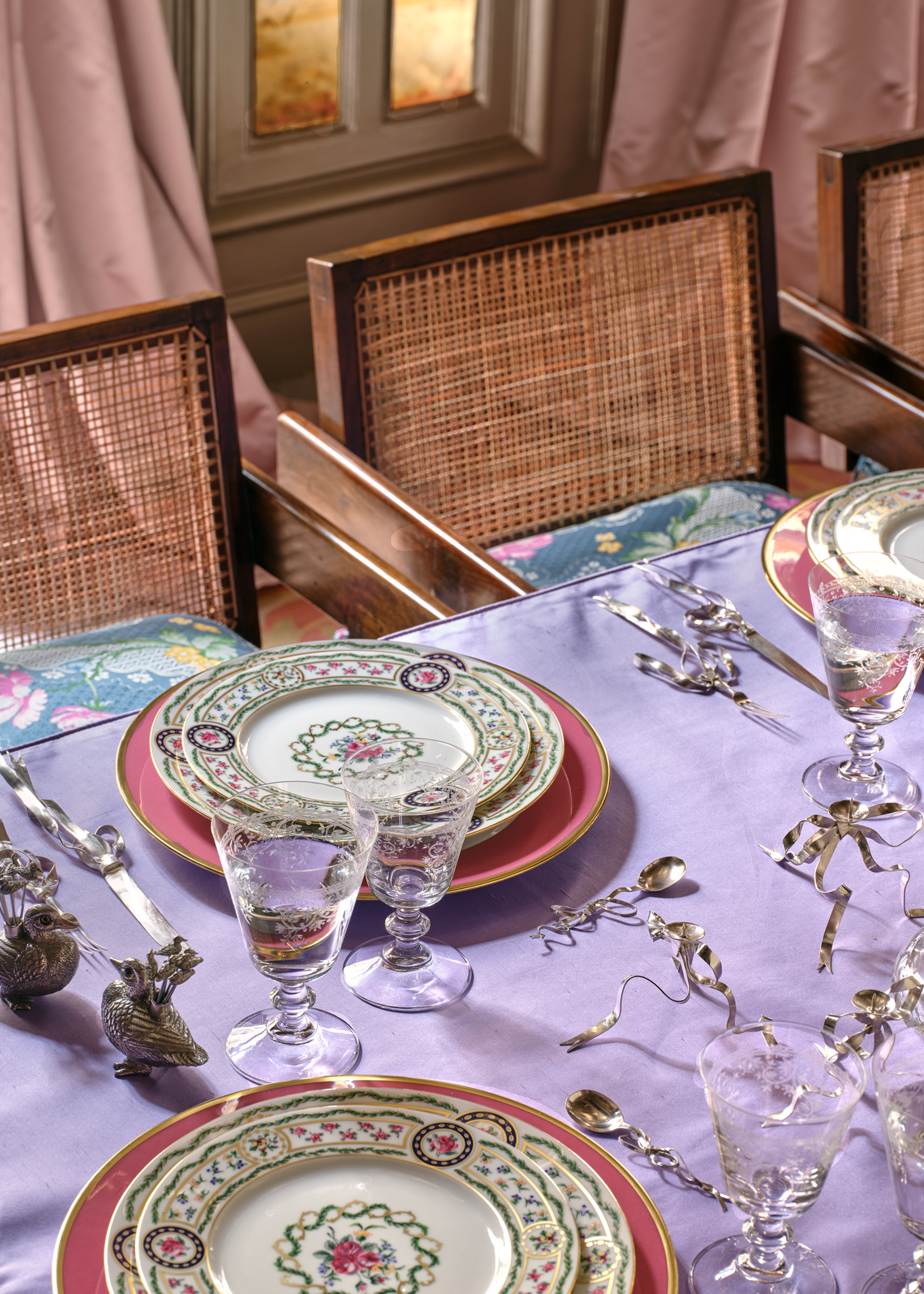 A table set for a meal with a purple table cloth, pink charger plates, and patterened plates on top with chrome bow decor on the table and rattan dining chairs