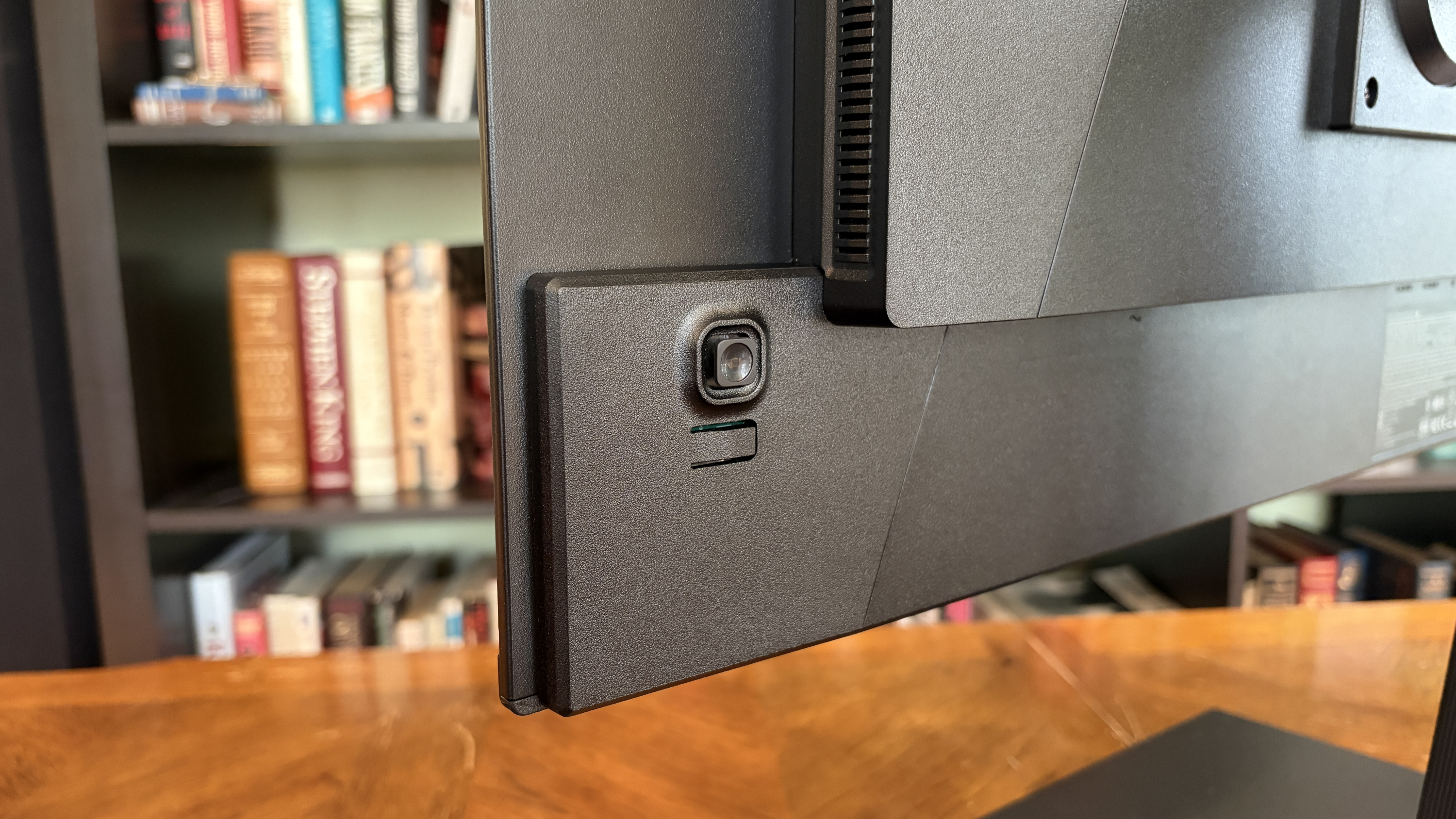 The close-up image shows the bottom corner of a black computer monitor with a camera. A bookshelf filled with books is in the blurred background.