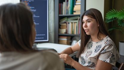 Upskilling concept image showing two female office IT workers discussing no-code tools with binary code pictured on desktop monitor.