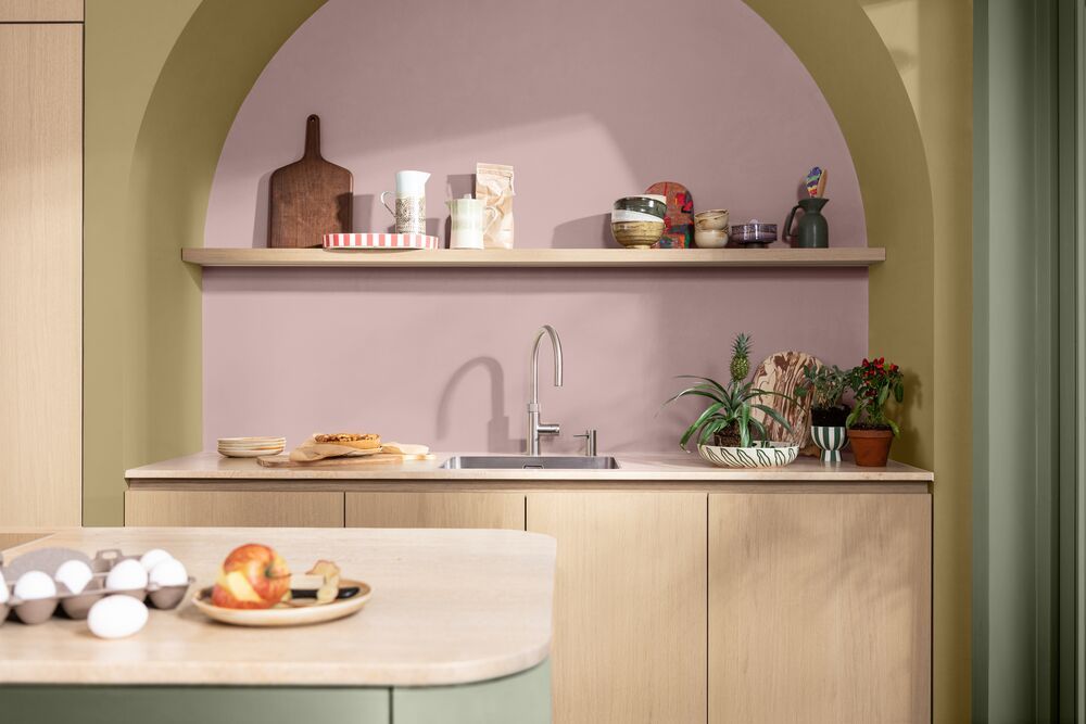 A kitchen with wooden cabinetry and an island with a sage green base. The sink on the back wall has a backsplash painted light pink and yelow with sage green trim around the room.