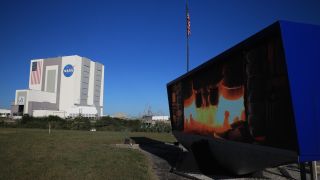 A grassy field leads to a large cube building stands against a blue sky in the background. In the foreground on the right, a blue screen showing the business end of a rocket as it launches.