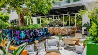 A street art-informed hotel courtyard features multiple plants, soft and rattan seating, a mosaic bar counter, and plenty of natural sunlight.