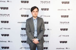 Josh Brener standing in front of a white backing board wearing a grey suit.