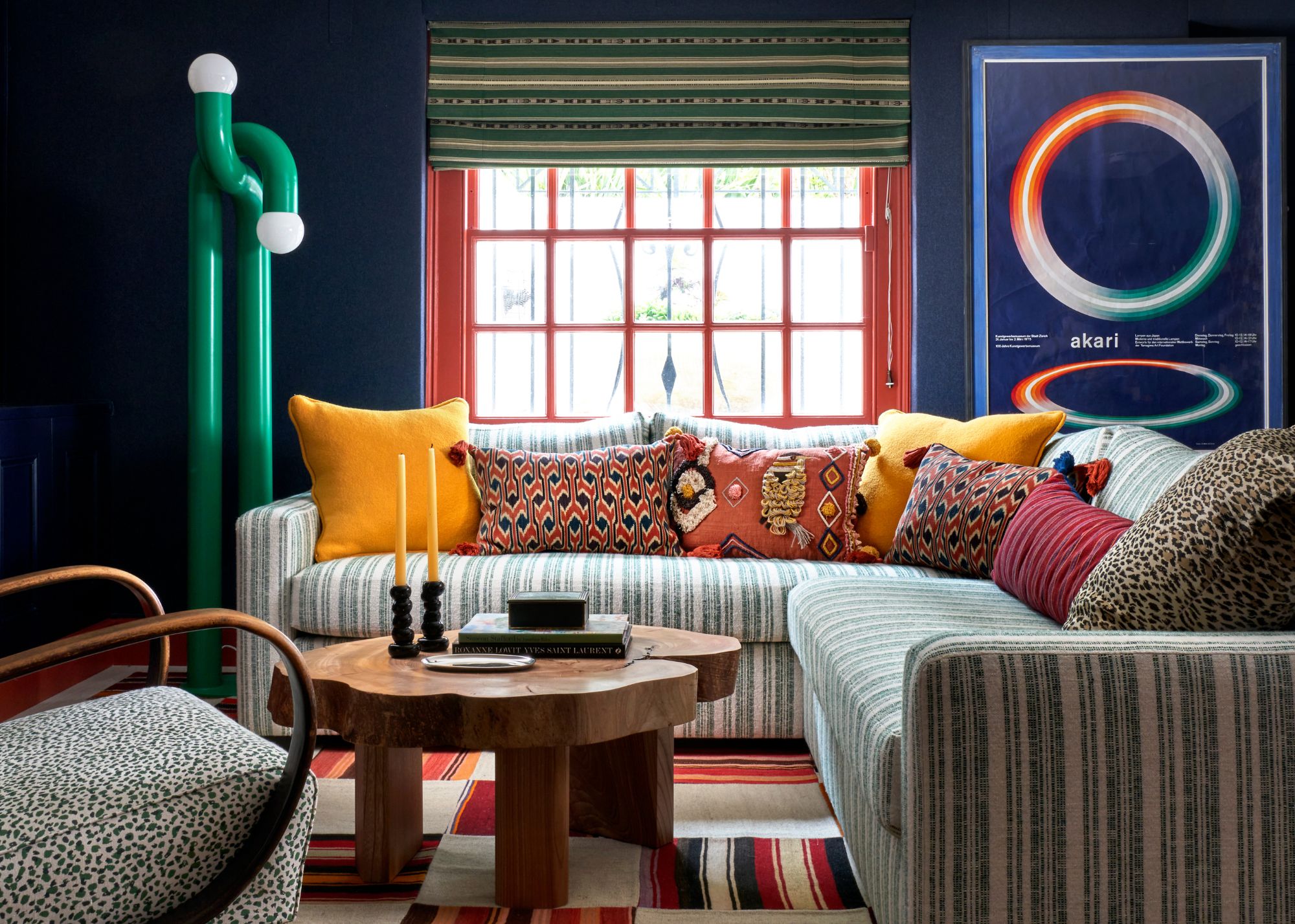 A living room with dark blue walls, red window trim, a green striped corner sofa, a wooden coffee table, a red and cream checkered rug, and an abstract and minimal green lamp.