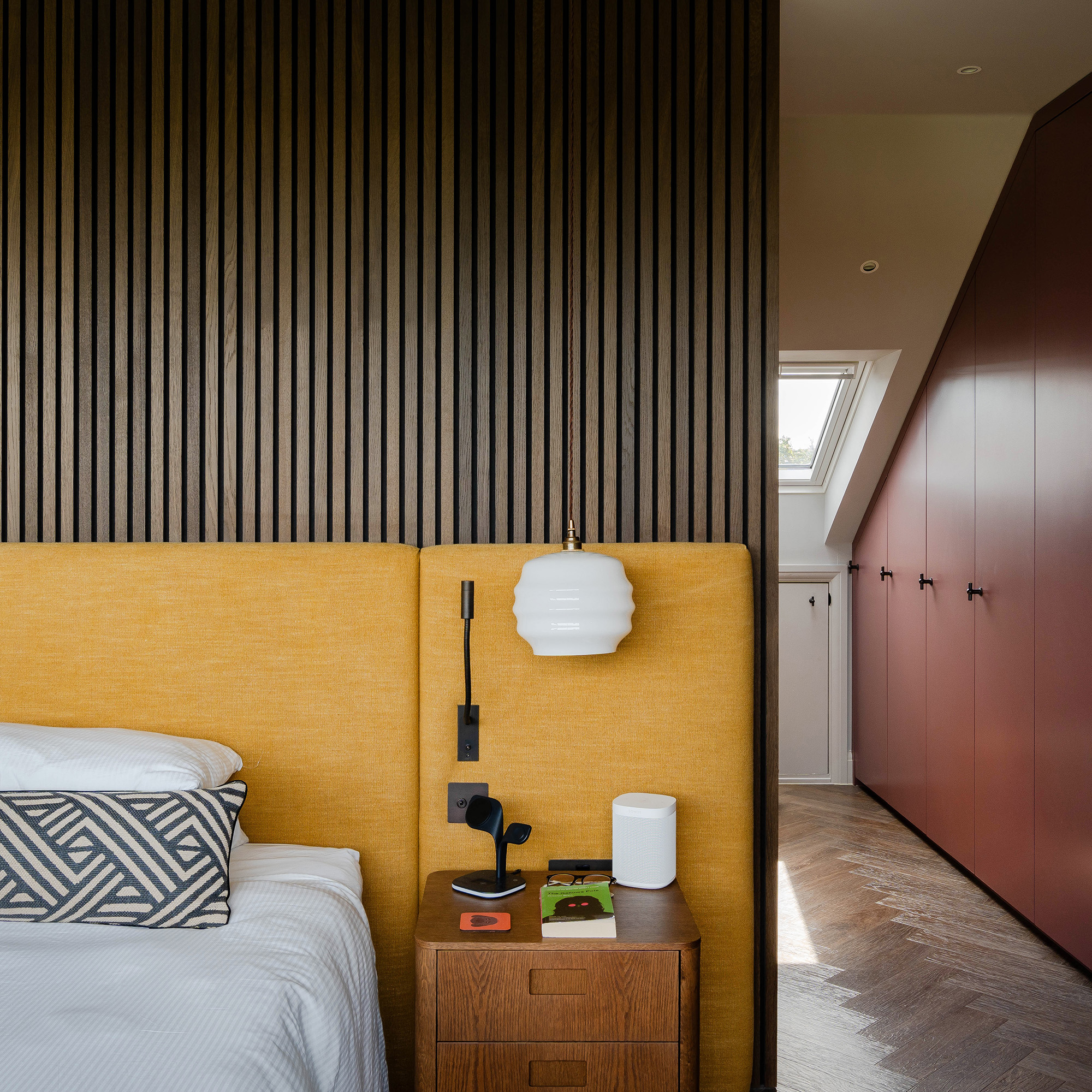 bedroom with wood clad walls and upholstered yellow headboard leading onto a dressing room with red built in furniture
