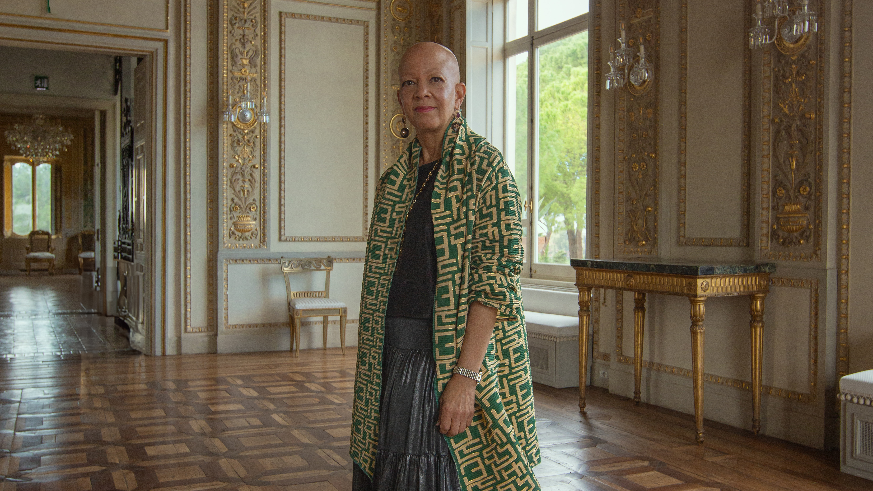 Shelia Bridges looking to camera wearing a green coat in a grand palazzo
