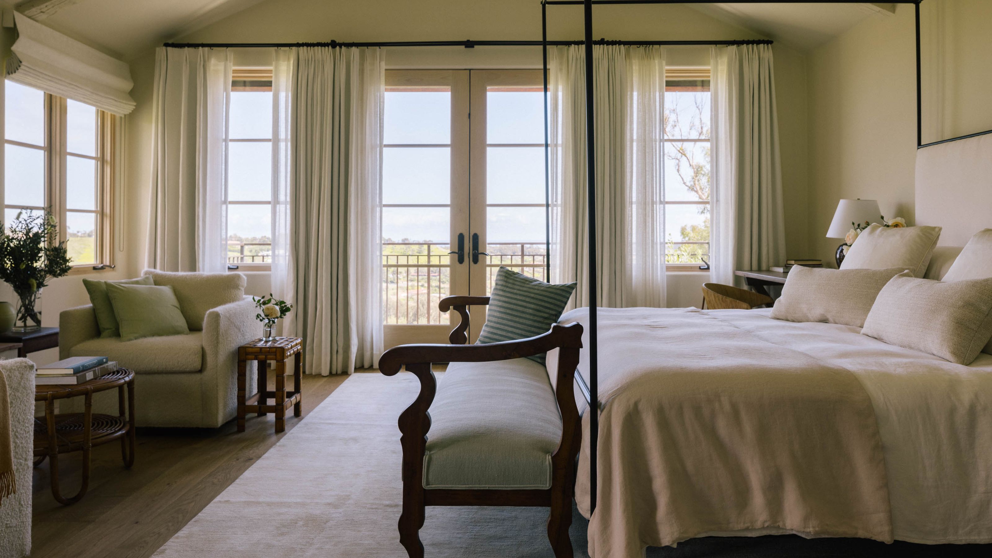 a large neutral primary bedroom with large french doors, drapes, a wrought iron four poster bed, wooden foot of bed bench and a small sitting area