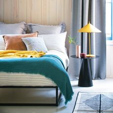 A bedroom with a bed dressed in multicoloured bedlinen and a bedside table holding a lamp with a cone-shpaed shade