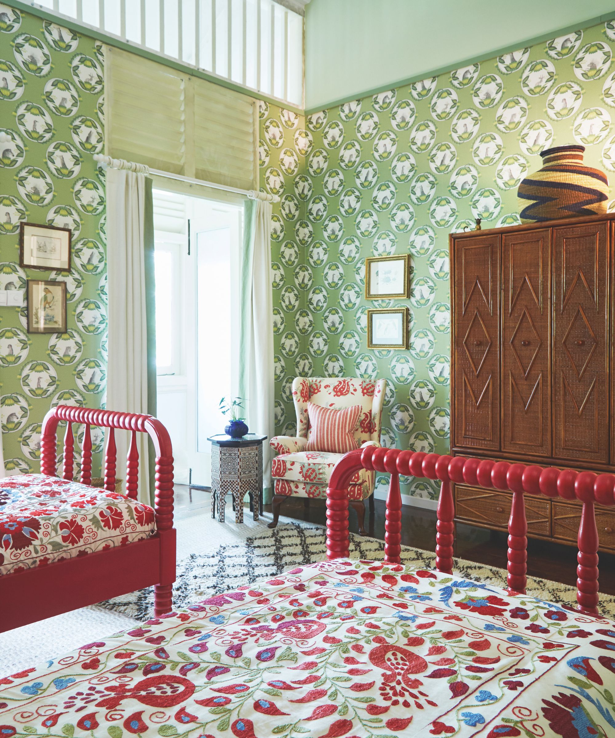 A maximalist bedroom with two red single beds, with bright patterned bedding, a black and white patterned rug, patterned upholstered chair, green patterned wallpaper, wooden closet, and large window with sheer white curtains.