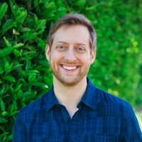 Brandon is a white man with a dark blue checkered collared shirt on, standing outside against a tall green privet hedge