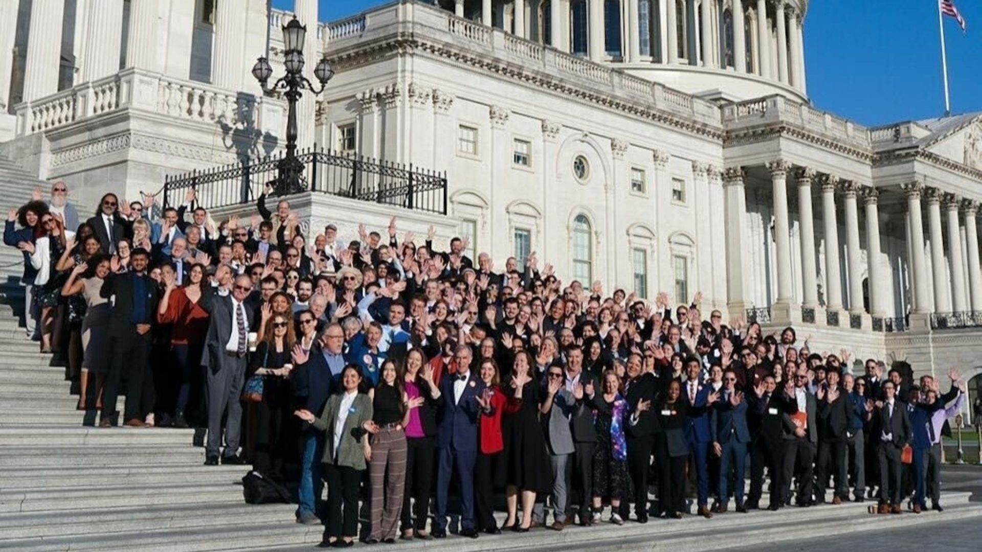 A large group of people stand on the steps of the U.S. Capitol in bright sunlight.