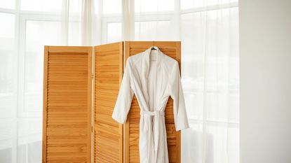 A white bathrobe hanging from a coat hangar on the back of a wooden screen divider. In front of a large bay window covered with a sheer white curtain