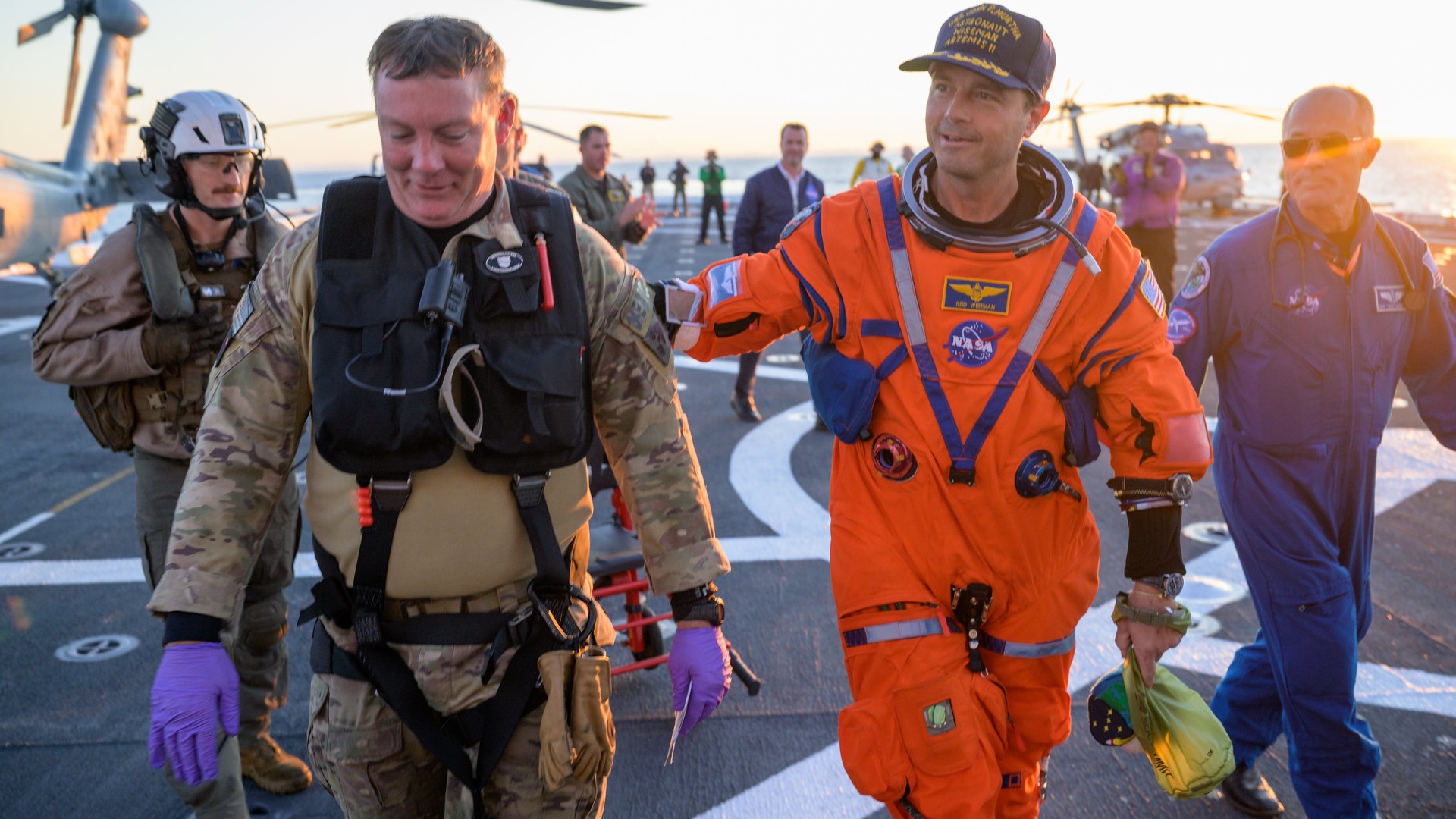 a man in an orange flight suit smiles while carrying a small stuffed moon