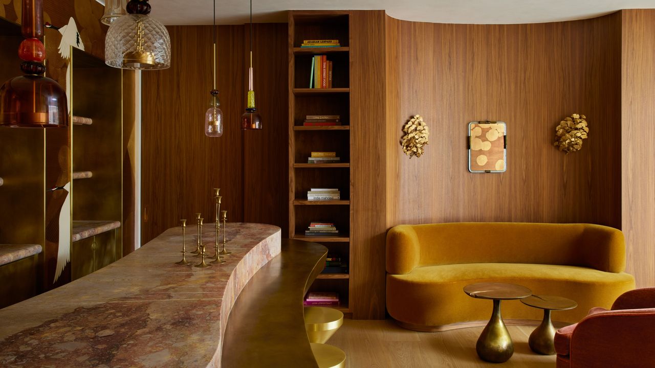 Image of a living room that merges into an at-home bar. The walls are made of wood and have a slight curve to them. There is a curved ochre yellow sofa with a gold nesting coffee table in front of it. The bar is made of a pink marble material with a gold off-shoot and gold bar stools. 