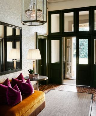 entryway with textured wallpaper, dark green mirror and wood work, tiled floor, turmeric seating with plum pillows, rug, side table and lamp, chandelier