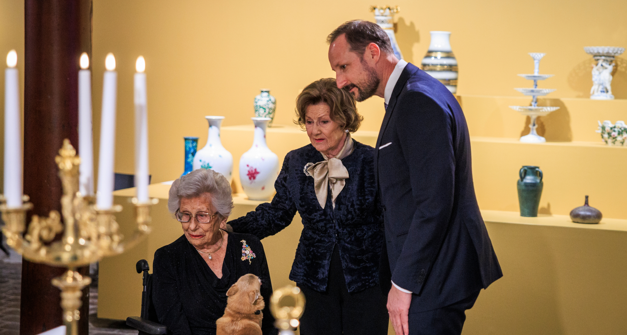 King Haakon, Queen Sonja and Princess Astrid, sitting in a wheelchair with her dog, viewing an exhibition 
