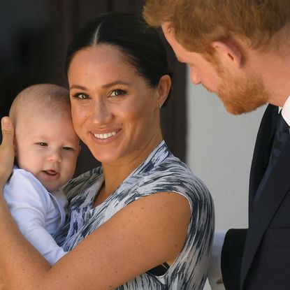 Meghan Markle wearing a blue patterned dress holding baby Prince Archie and smiling while Prince Harry, wearing a suit, looks on