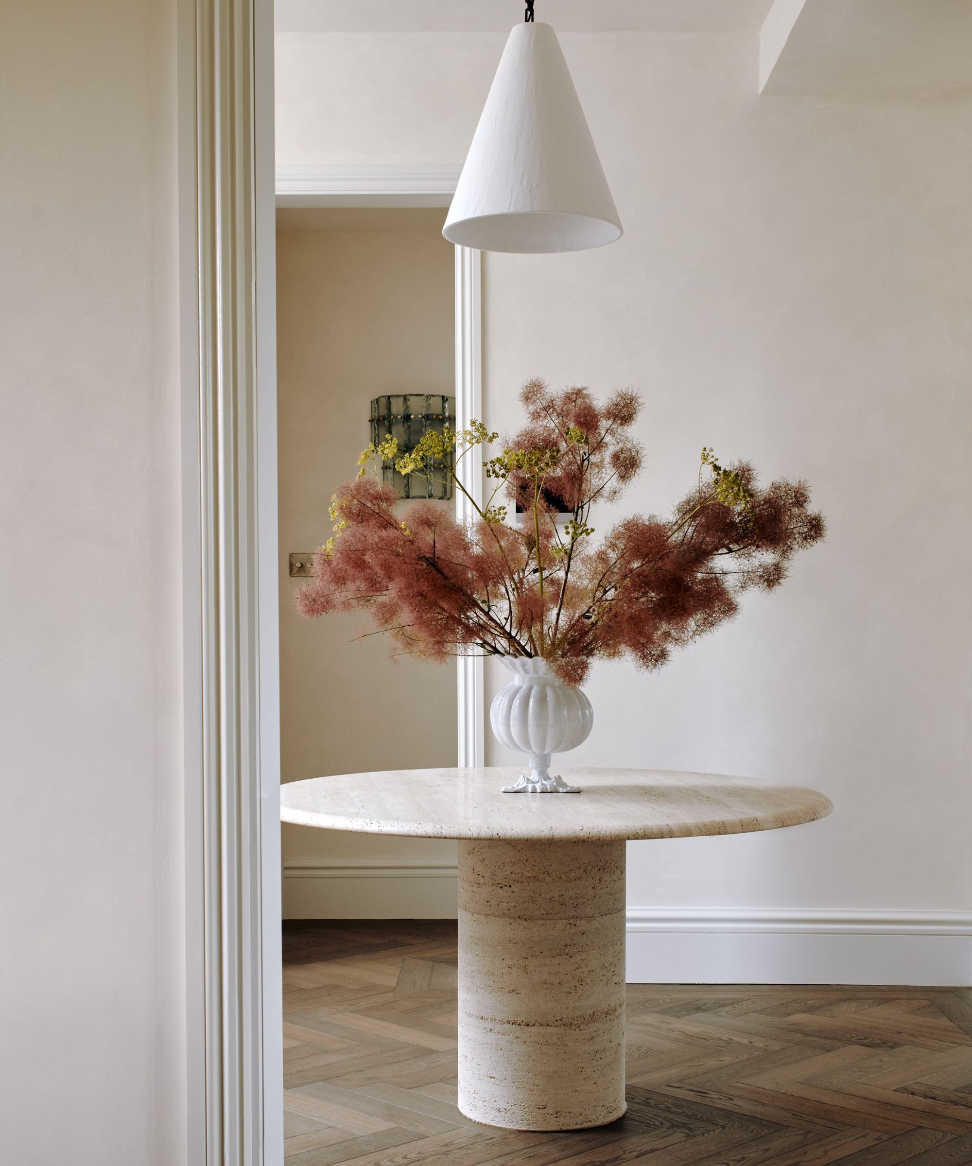 Round marble table in entance, decorated with large vase of pink flowers, white painted walls and dark wood flooring
