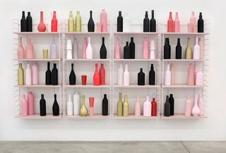 View of ’Early Retirement’ by Matthew Brannon featuring a pink, wall-mounted shelving unit with bottles in different colours on a light coloured wall in a space with grey flooring