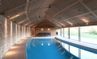 Contour House, swimming pool in the centre, stone walkway around the pool, brick wall along the left and glass windows along the right, wooden slat design ceiling with metal suuport frame, spot lights, end wall has wooden slat design with two high wall lights attached , four lounger seating, daytime outside view of the landscape through the windows