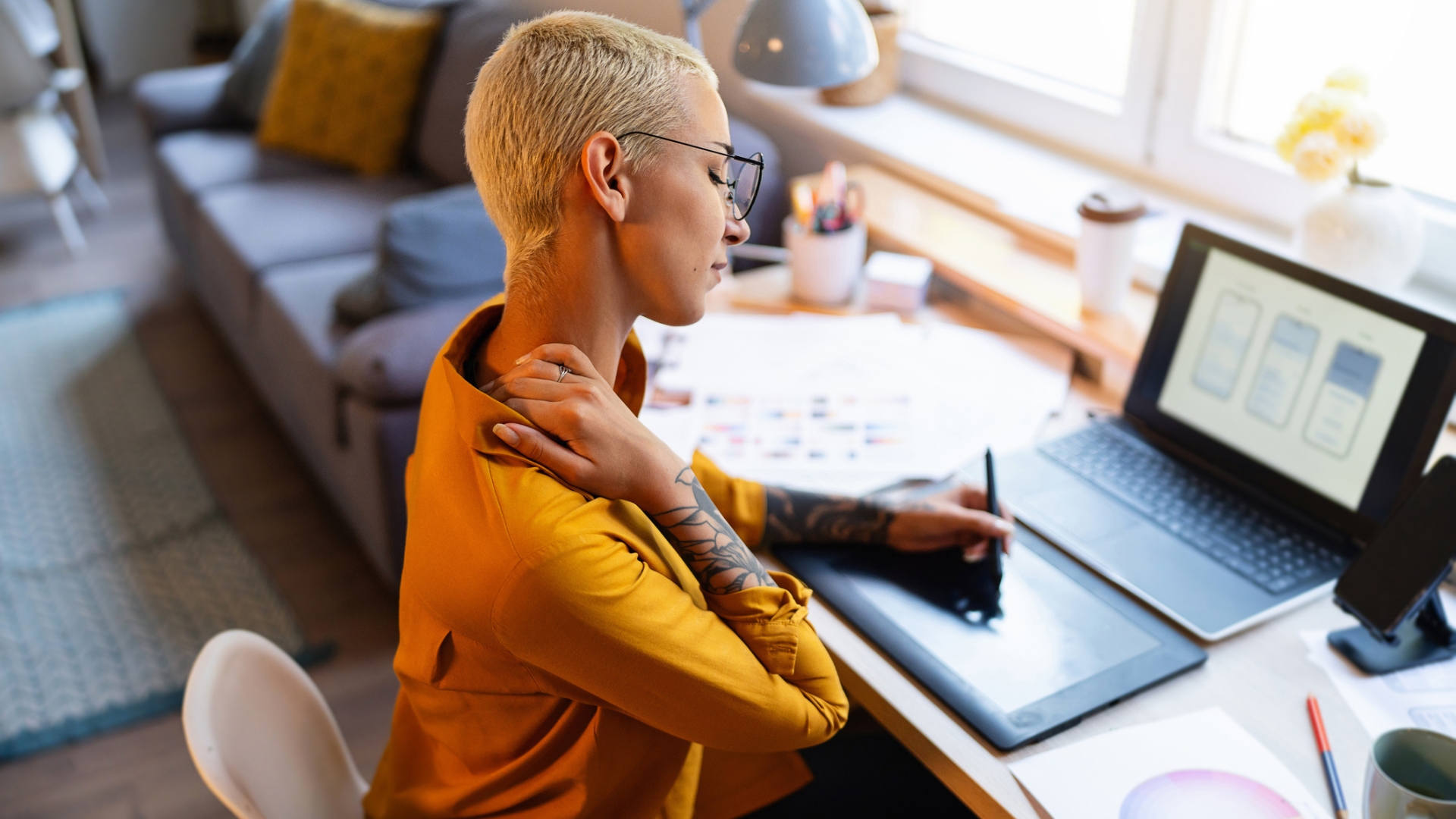 woman in a yellow shirt at a laptop and drawing device holding a pen in one hand and holding the side of her neck with the other. 