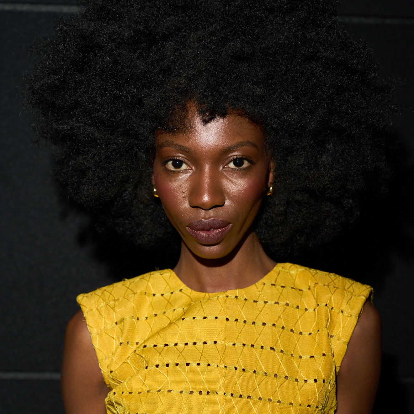 A model wearing a yellow top and her dark curly hair styled with micro bangs.