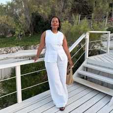 A woman wearing a linen vest and pant set with a raffia handbag, black sandals, and gold hoops.