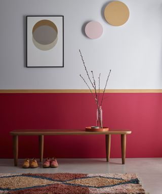 hallway with lower half of wall painted red topped with a thin yellow border and soft grey wall above, wooden bench, rug and artwork on walls