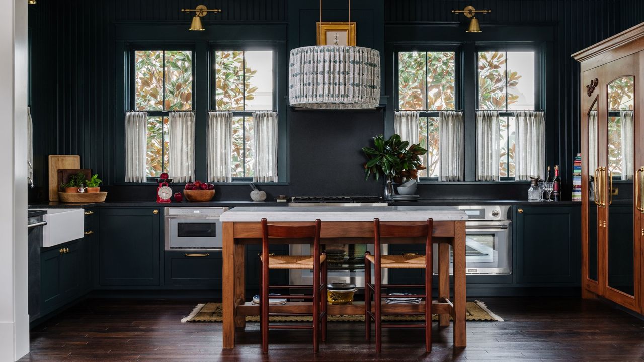 dark green kitchen painted in benjamin moore&#039;s salamander shade, with timber freestanding island bench, a pendant light hanging overhead, cafe curtains on windows, and a freestanding timber cabinet and butler&#039;s sink