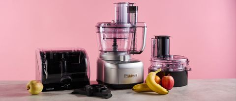 The Cuisinart Expert Prep Pro food processor pictured against a pink background. The accessories storage case is pictured to its left, and the dicing accessory kit is pictured on its right. The stone-effect tabletop that it sits on has apples and bananas on.