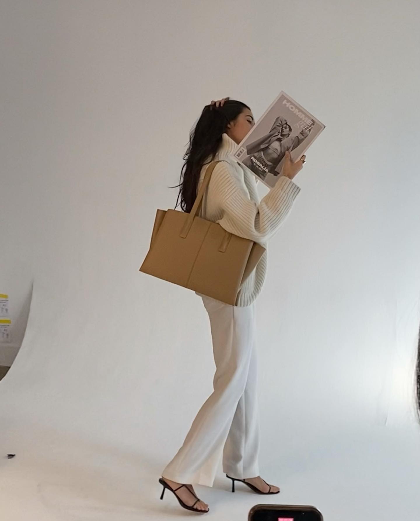 Woman in front of white backdrop wearing all white, holding newspaper, and wearing an oversize Freja tote in a khaki color on her shoulder.