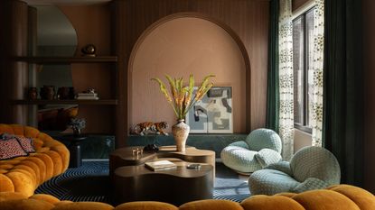 Abstract and colorful living room with curved coffee table, large potted plant, orange, round, padded sofa, and two soft chairs. A wooden archway and shelves with miscellaneous ornaments are pictured in the background.