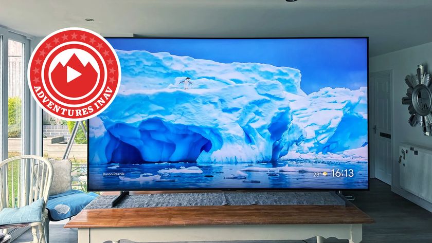 The 98-inch TCL C7K pictured on a wooden table. In the corner of the photo is a red logo that says &#039;Adventures in AV&#039;.