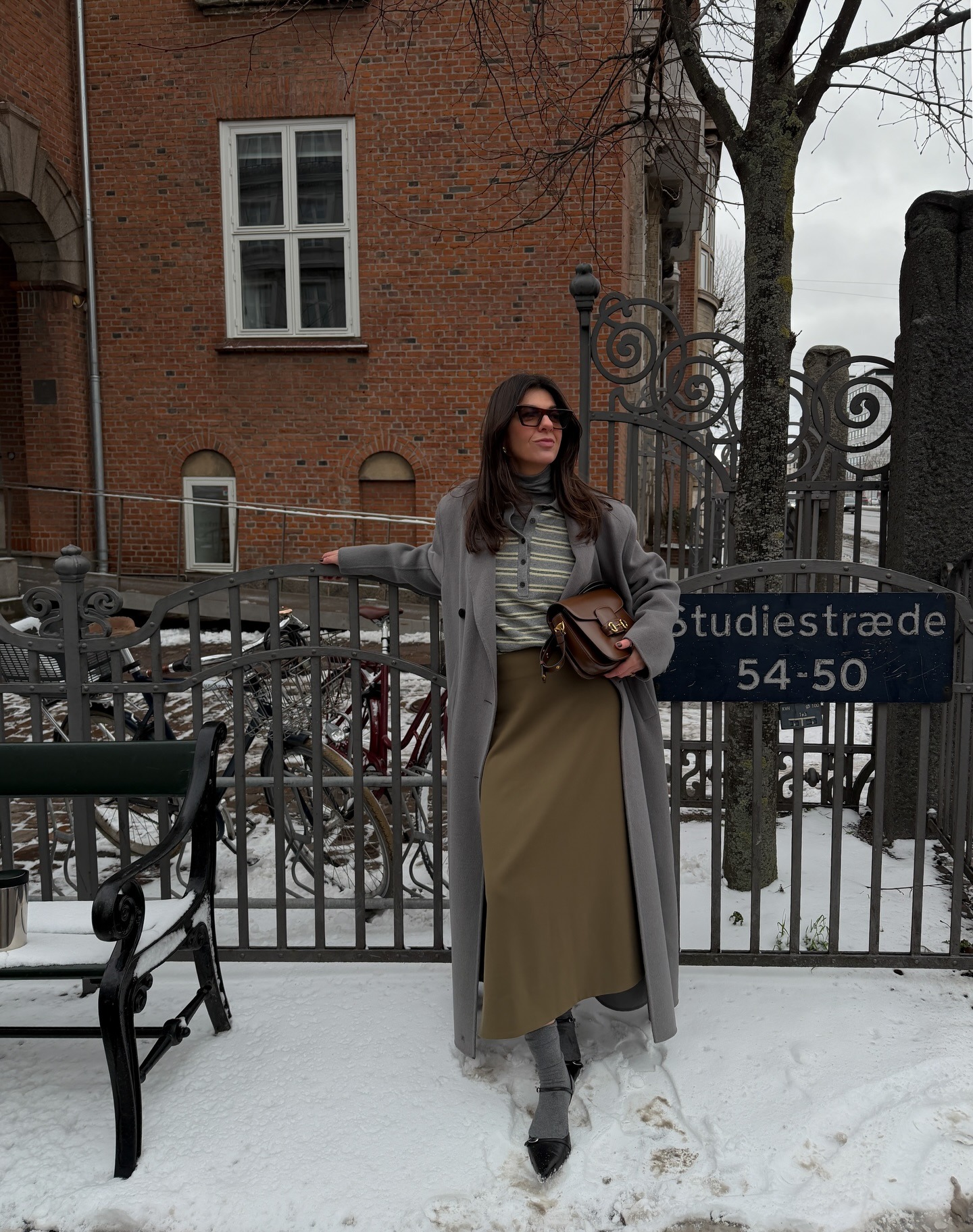 Fashion person francesca saffari show whats to wear in February 2026. She wear a grey turtle neck layered underneath a blue grey and white striped polo. On top she wears a grey coat and she finishes the look with a green maxi skirt. In her hand she carries the Gucci Horsebit bag in brown. This image was recently taken from her Instagram.