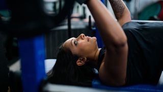Poorna Bell underneath a barbell, lying back on a bench in the gym, about to complete a bench press