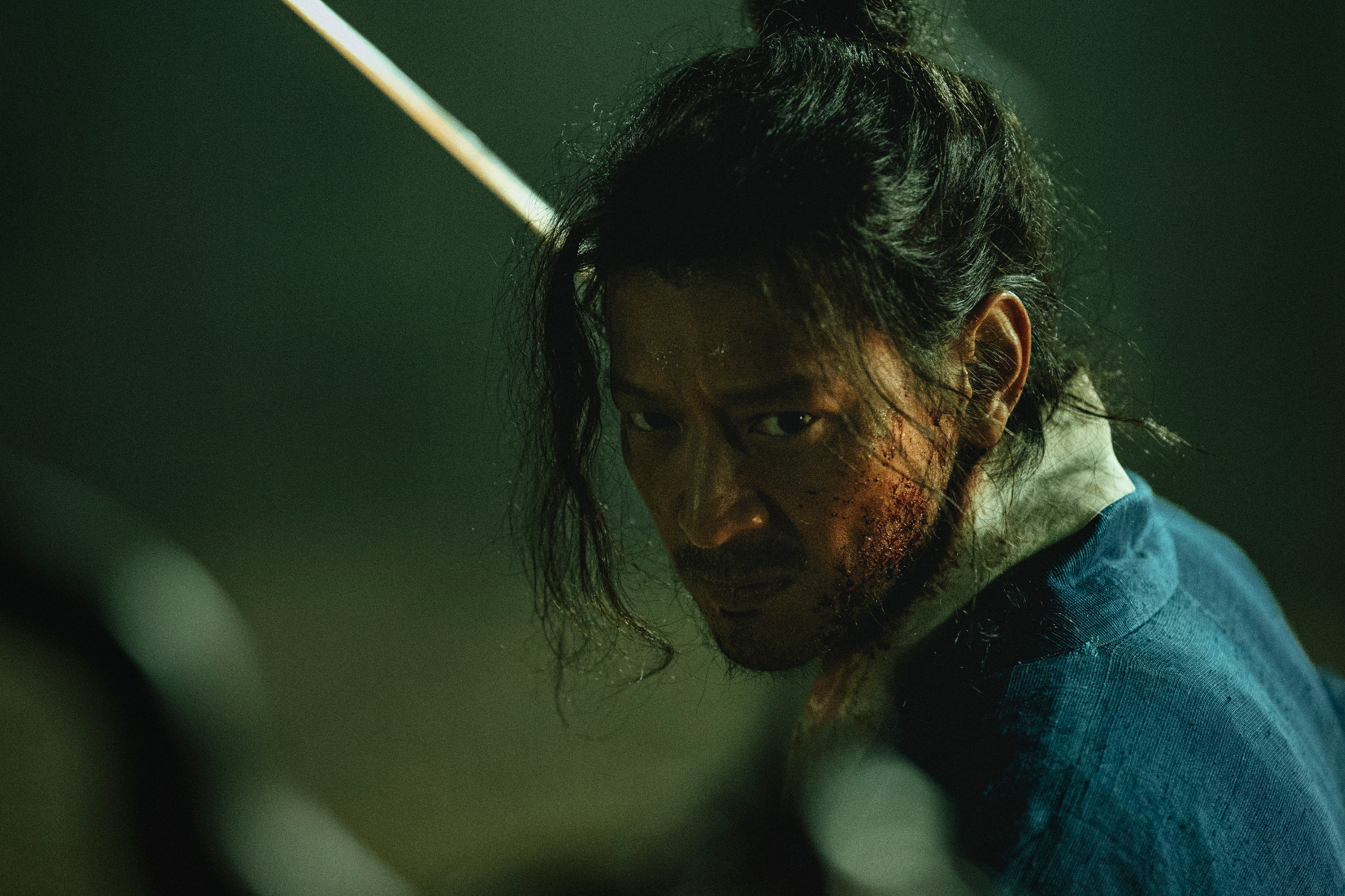 A man is covered in sweat and grime as he wears a traditional Korean outfit. A sword is seen in the blurred background, in the Korean movie 'Uprising.'