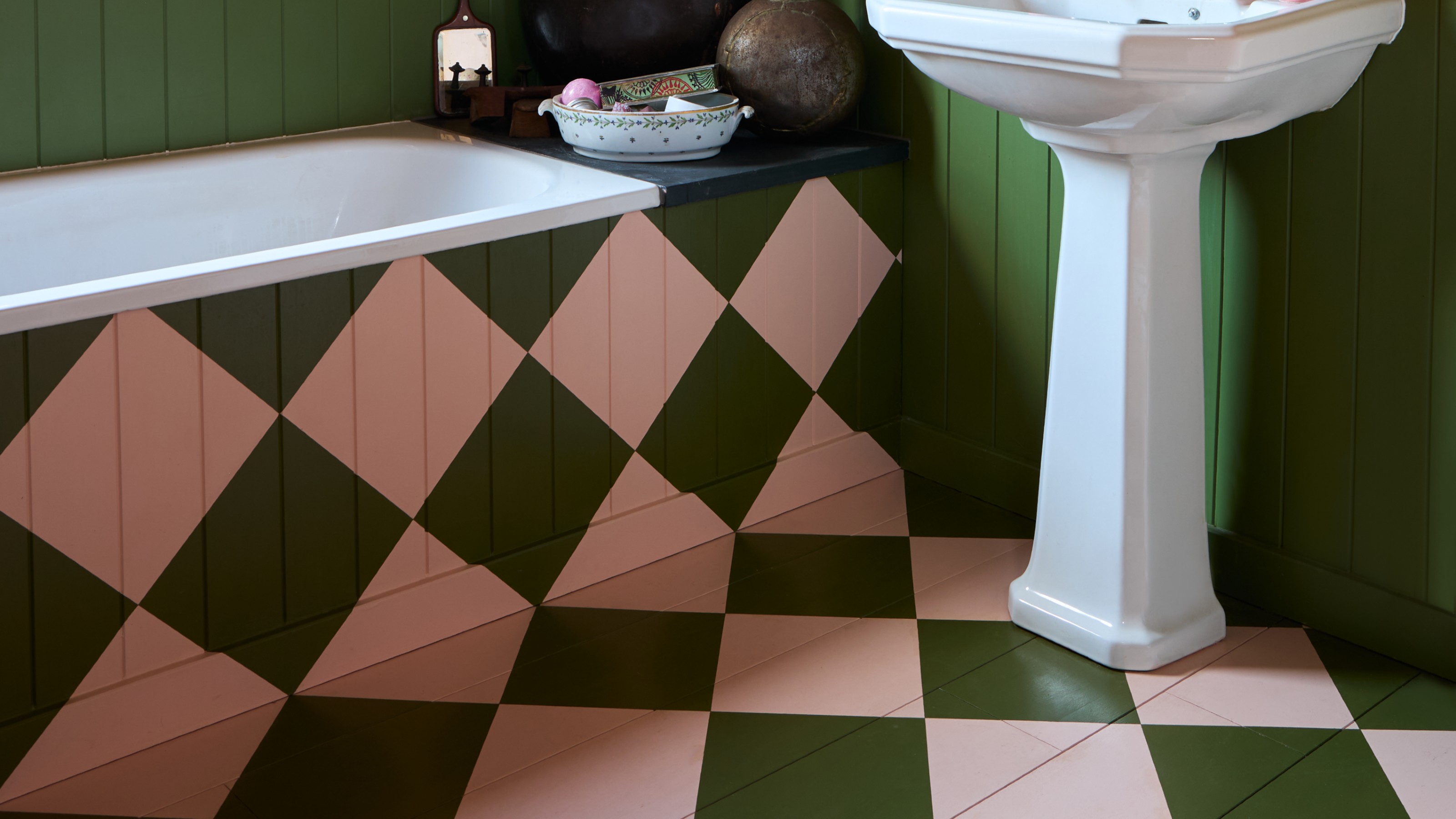 A bathroom with wooden floors and bath panelling painted in a chequerboard pattern in soft pink and olive green shades