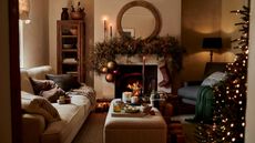 A warm Christmas living room, with lime washed walls and a fireplace. A large wreath over the mantel with a hanging stocking. The edge of a tree visible from the edge of the doorway.