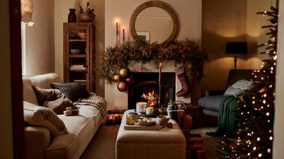 A warm Christmas living room, with lime washed walls and a fireplace. A large wreath over the mantel with a hanging stocking. The edge of a tree visible from the edge of the doorway.