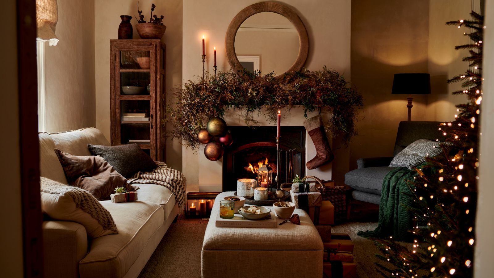 A warm Christmas living room, with lime washed walls and a fireplace. A large wreath over the mantel with a hanging stocking. The edge of a tree visible from the edge of the doorway.