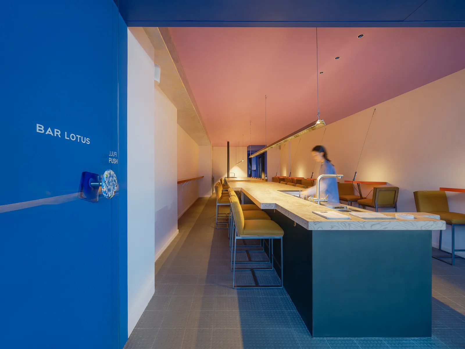 A blue-tinted, minimalist bar features a long, frills-free bar with yellow velvet mid-century modern stools behind which a female bartender is working, pink ceilings, and one elongated neon light running throughout it.