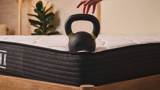 A 35lb kettlebell balances on the corner of the Plank Firm mattress, Extra Firm side, with a hand hovering over the top