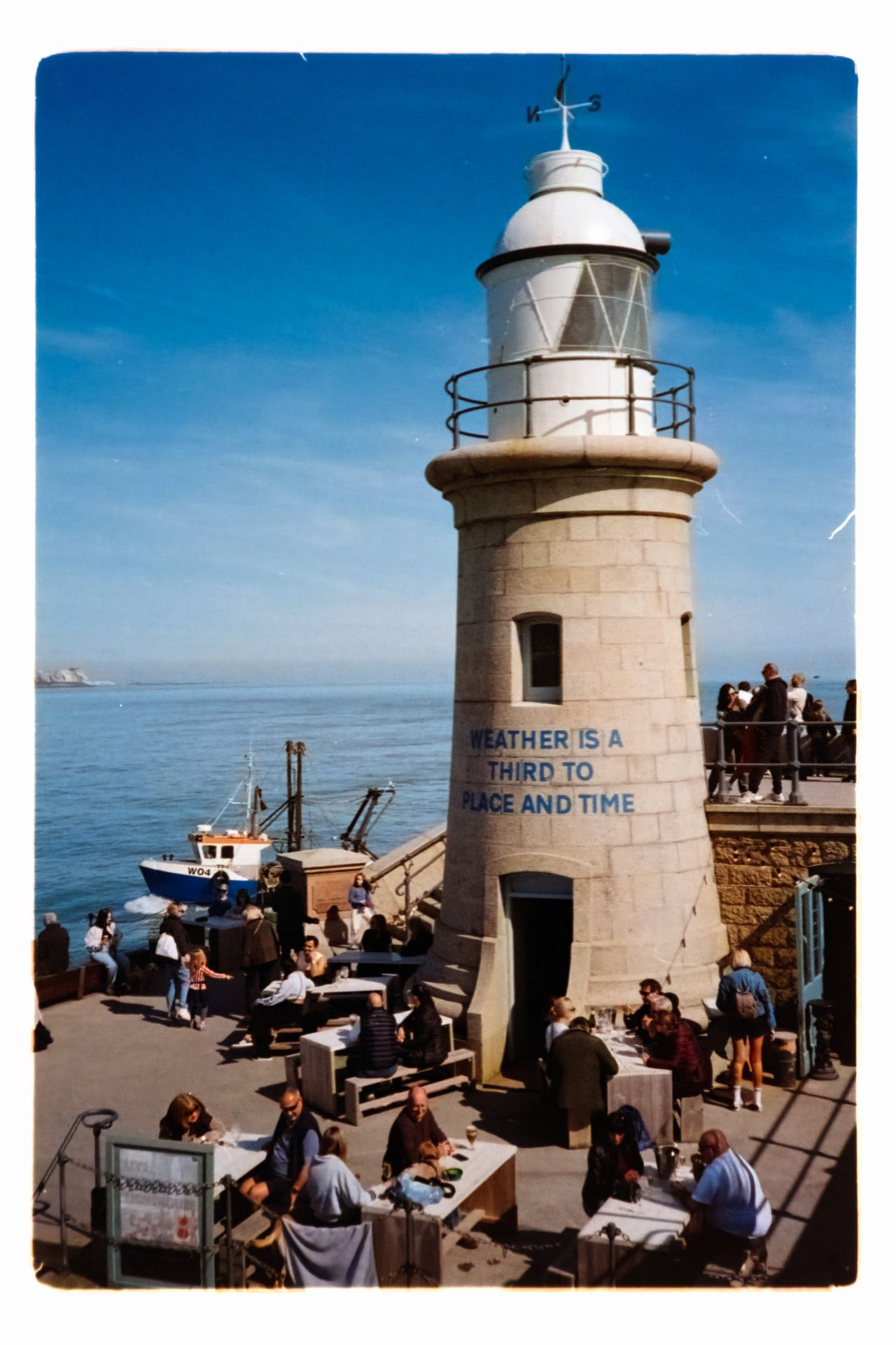 Scanned color film photograph of a small lighthouse beside the sea, surrounded by people dining and walking on the waterfront.