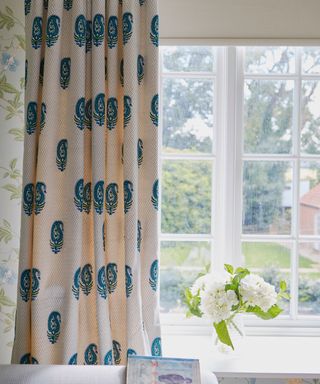 A closeup of a window and floral wallpaper, dress curtain and curtain pole and roman blinds.