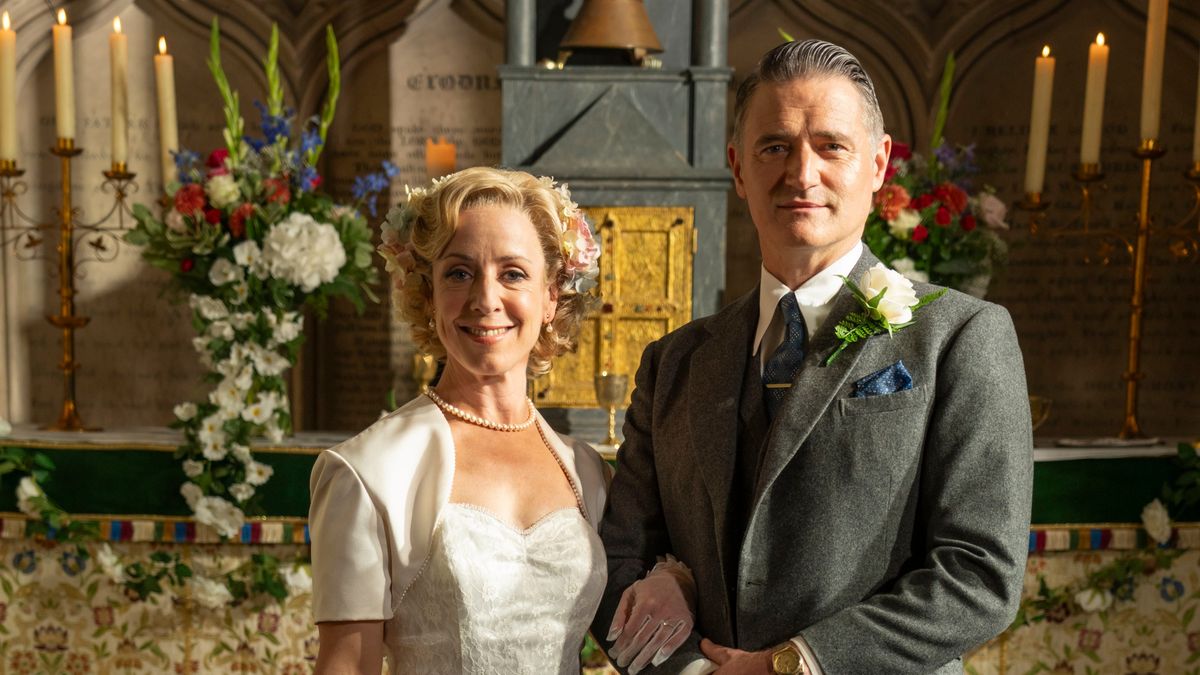 Mrs Devine (Claudie Blakley) and Chief Inspector Sullivan (Tom Chambers) stand at the altar in their wedding outfits, facing the camera. She is in an ivory dress with a bolero-style jacket oer the top. He is wearing a grey morning suit with a white flower in his lapel. The altar behind them has a beautiful display of mostly white flowers with some red, orange and blue as well.