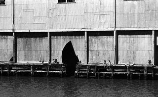 Black and white image - people sitting on a jetty in front of a large wooden-like building.