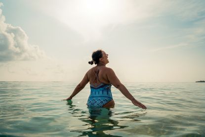 A woman in a patterned blue one-piece swimsuit stands in the sea smiling with her hands touching the water.