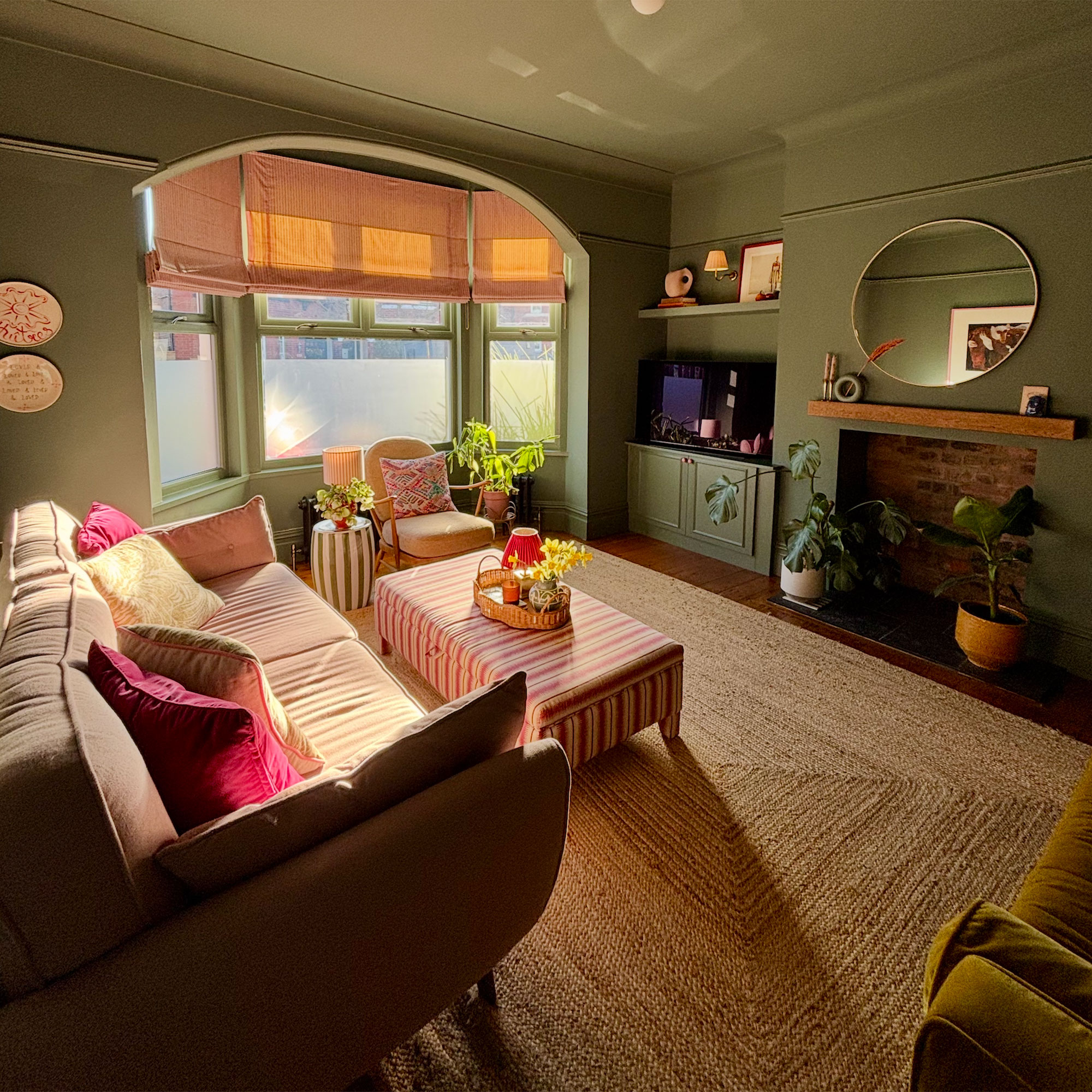 Green living room with jute rug, pink blinds in bay window and striped upholstered ottoman used as coffee table