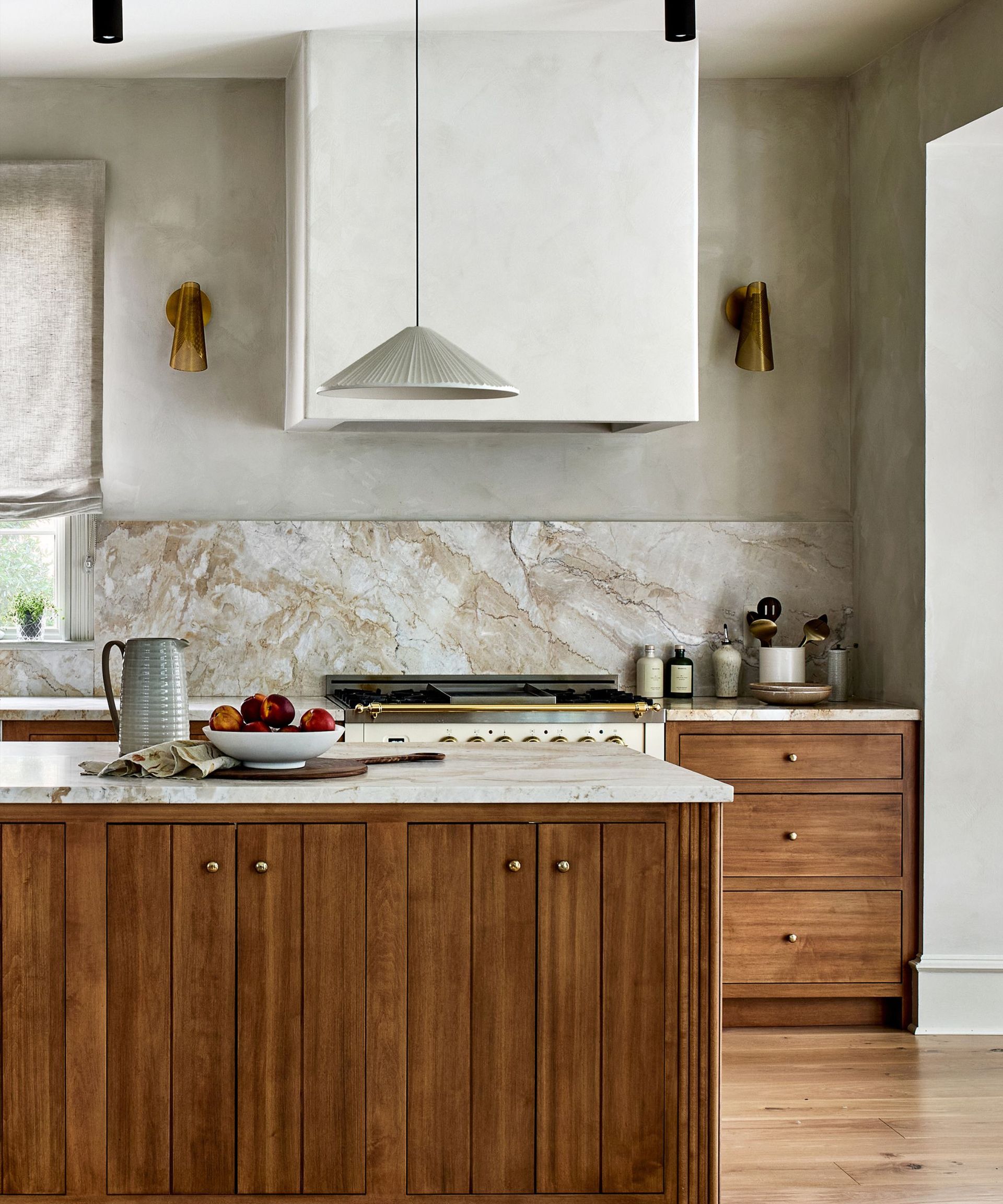 A kitchen with a wooden island, wood cabinets, marble countertops, limewash walls, a plaster range hood, and layered lighting